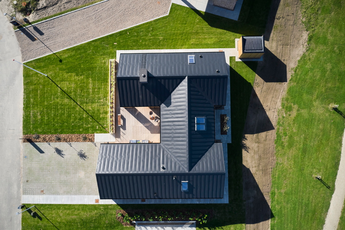 H-hus fotograferet fra luften med mørkt tag, ovenlysvinduer, centralt terrassemiljø, flisebelagt indkørsel og grøn have – inspiration til moderne nybyg og husdrømme.