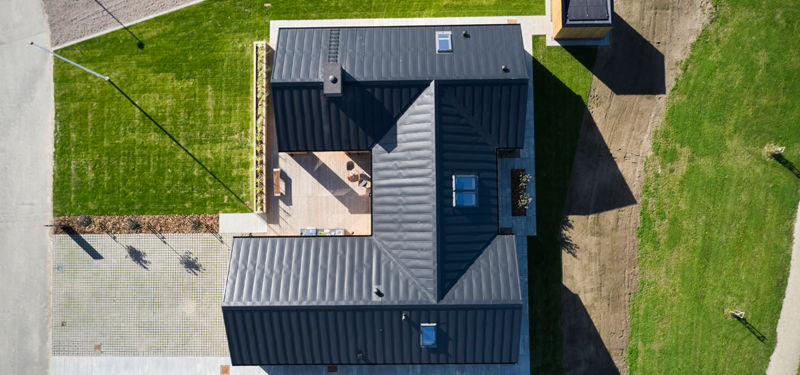 H-hus fotograferet fra luften med mørkt tag, ovenlysvinduer, centralt terrassemiljø, flisebelagt indkørsel og grøn have – inspiration til moderne nybyg og husdrømme.