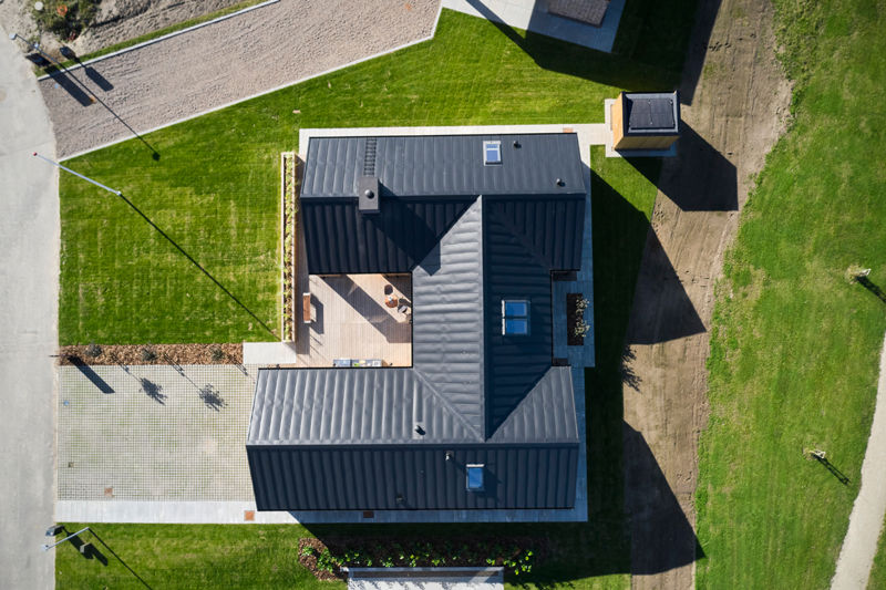 H-hus fotograferet fra luften med mørkt tag, ovenlysvinduer, centralt terrassemiljø, flisebelagt indkørsel og grøn have – inspiration til moderne nybyg og husdrømme.