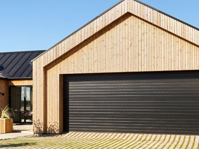 Udvendig garage med stor sort garageport, lodret træbeklædning, sort tag, bred indkørsel med græsarmering og overdækket terrasse ved siden af – inspiration til garage i nybyggeri, carportløsninger og moderne husdrømme fra eurodan-huse.