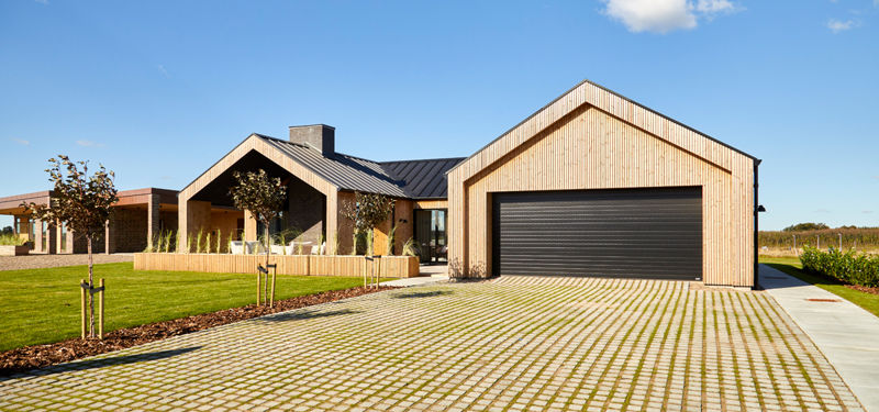 Udvendig garage med stor sort garageport, lodret træbeklædning, sort tag, bred indkørsel med græsarmering og overdækket terrasse ved siden af – inspiration til garage i nybyggeri, carportløsninger og moderne husdrømme fra eurodan-huse.