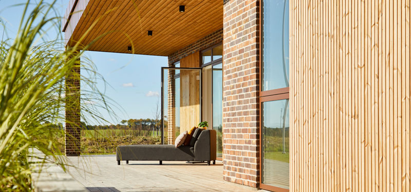 Overdækket træterrasse i udstillingshus fra eurodan-huse med træfacade, murstenssøjle, loungemøbler og store glaspartier til moderne nybyggeri og husbyggeri.