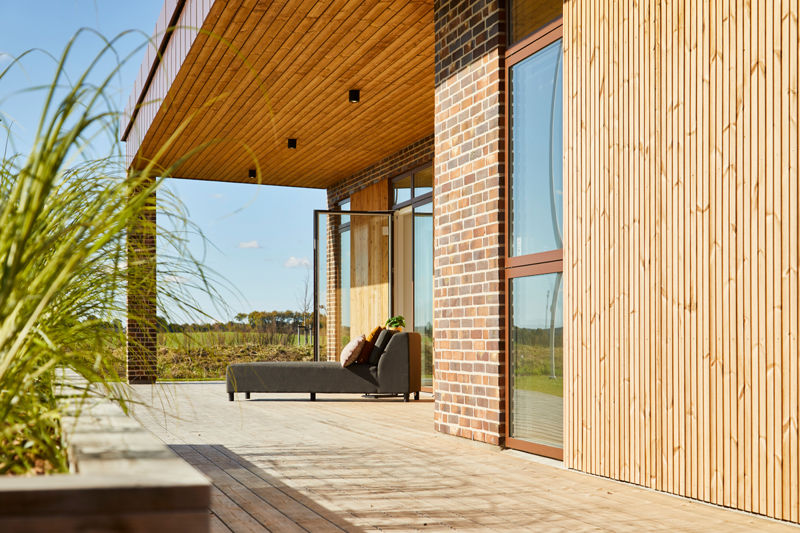 Overdækket træterrasse i udstillingshus fra eurodan-huse med træfacade, murstenssøjle, loungemøbler og store glaspartier til moderne nybyggeri og husbyggeri.