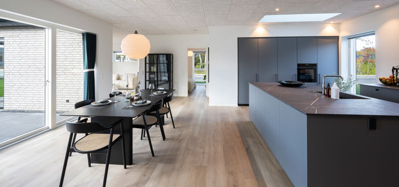 Lyst køkken-alrum med mørkt køkken, spisebord og store glaspartier i et moderne drømmehus.