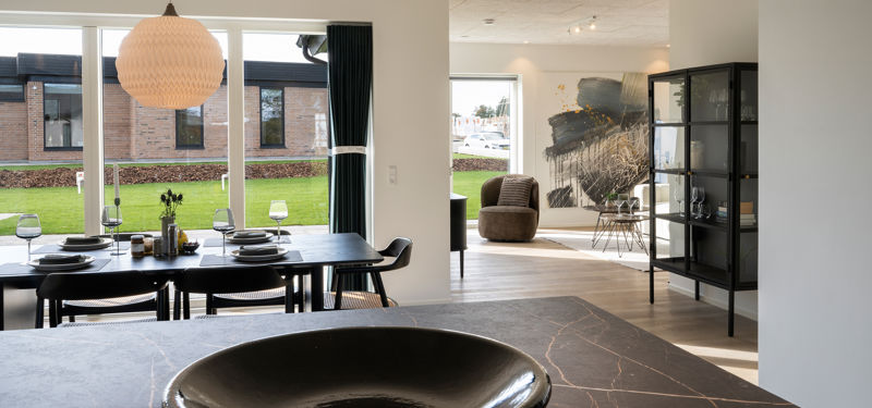 Udsigt fra køkkenet mod spiseplads og opholdsstue med store vinduer og naturligt lys i det nye hus.