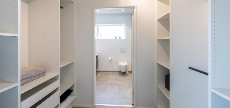 Walk-in closet med åbne hvide garderobemoduler, hylder, skuffer, sorte bøjlestænger, tekstiler på hylderne og direkte adgang til badeværelse i baggrunden – inspiration til funktionel opbevaring, nybyggeri og gennemtænkte husdrømme fra eurodan-huse.