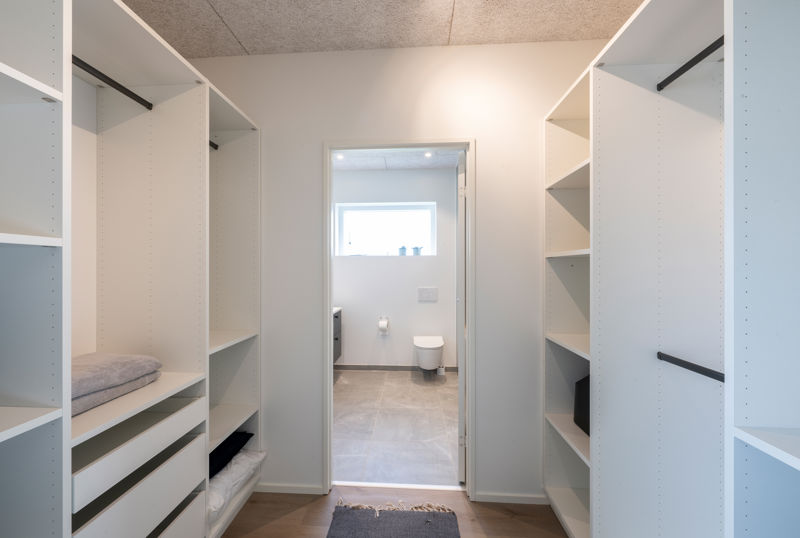 Walk-in closet med åbne hvide garderobemoduler, hylder, skuffer, sorte bøjlestænger, tekstiler på hylderne og direkte adgang til badeværelse i baggrunden – inspiration til funktionel opbevaring, nybyggeri og gennemtænkte husdrømme fra eurodan-huse.
