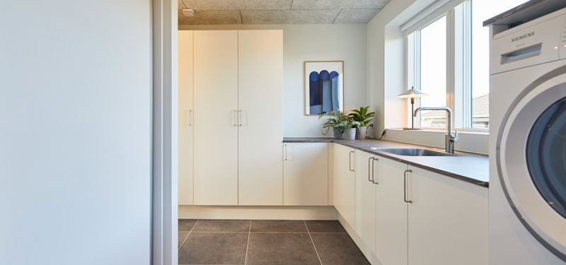 Bryggers fra eurodan-huse med mørkt klinkegulv, hvide højskabe, vaskemaskine og tørretumbler, bordplade langs vinduesvæggen, integreret vask og plante ved vinduet – inspiration til nybyggeri med god opbevaring, praktisk vaskerum og funktionelt husbyggeri.
