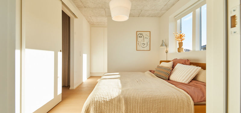 Soveværelse fra eurodan-huse med sollys over sengen, beige sengetæppe, rosa og mønstrede puder, vindue over sengegavlen, vægkunst, hvid skabsvæg og gardinåbning mod walk-in-område – inspiration til lyst nybyggeri, husbyggeri og varme husdrømme.
