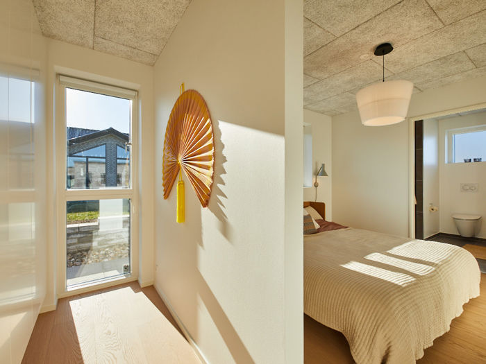 Soveværelse fra eurodan-huse med adskilt garderobeområde, højt vindue, dekorativ vægventilator, beige sengetæppe, rund loftlampe og kig til eget badeværelse med væghængt toilet – inspiration til nybyggeri med praktisk walk-in og gennemtænkte husdrømme.