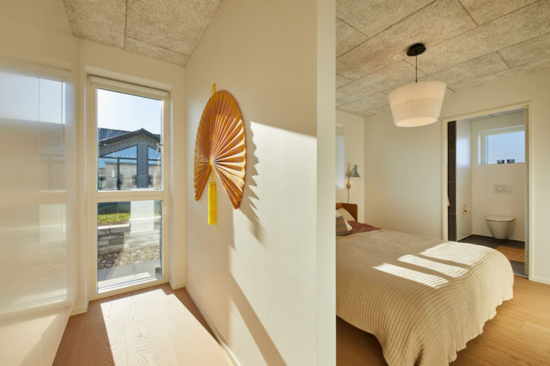 Soveværelse fra eurodan-huse med adskilt garderobeområde, højt vindue, dekorativ vægventilator, beige sengetæppe, rund loftlampe og kig til eget badeværelse med væghængt toilet – inspiration til nybyggeri med praktisk walk-in og gennemtænkte husdrømme.