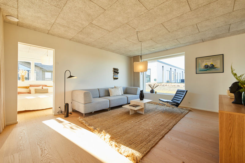 Stue fra eurodan-huse med lysegrå sofa, sort loungestol, jute-tæppe, hvidt sofabord, sort gulvlampe, stort vindue og åbent kig til soveværelse med dobbeltseng – inspiration til nybyggeri, husbyggeri og gennemtænkte opholdsrum.