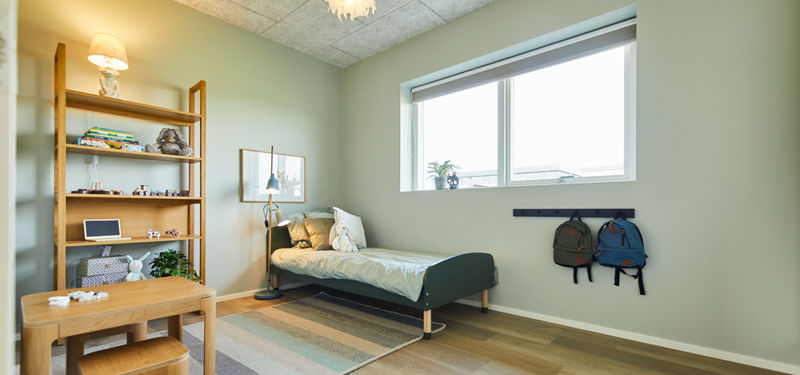 Børneværelse fra eurodan-huse med grønne vægge, enkeltseng, træreol, lille bord med skammel, gulvlampe, vægknager til tasker og bredt vindue – inspiration til funktionelle børneværelser i nybyggeri og husbyggeri.