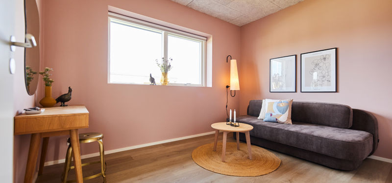 Værelse fra eurodan-huse med rosa vægge, mørk daybed, rundt sofabord på flettet tæppe, høj gulvlampe, lille skrivepult og stort vindue – inspiration til børneværelse, teenageværelse og personlige rum i moderne nybyggeri.