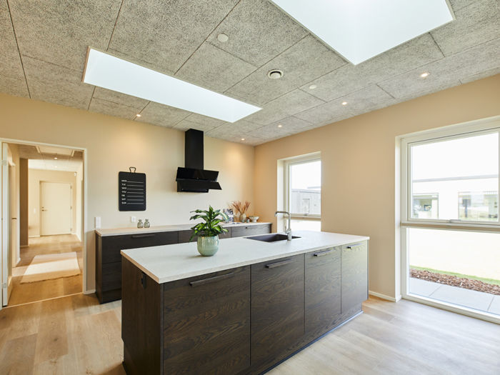 Moderne køkken i nybyg med mørke træfronter, stor køkkenø, lys bordplade, sort emhætte, ovenlys og brede vinduespartier – inspiration til køkkenet i drømmehuset.