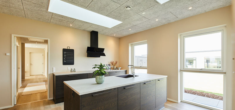Moderne køkken i nybyg med mørke træfronter, stor køkkenø, lys bordplade, sort emhætte, ovenlys og brede vinduespartier – inspiration til køkkenet i drømmehuset.