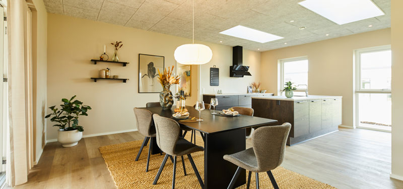 Lyst køkken-alrum i vinkelhus med mørkt spisebord, polstrede stole, køkkenø, store vinduer og ovenlys – inspiration til indretning af et moderne nybyg.