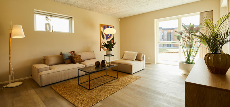 Stue fra eurodan-huse med beige sofa, matchende chaiselong, sort metalsofabord, stort vævet gulvtæppe, høj gulvlampe, grønne planter og stort vinduesparti med dagslys – inspiration til nybyggeri, husbyggeri og indbydende husdrømme.