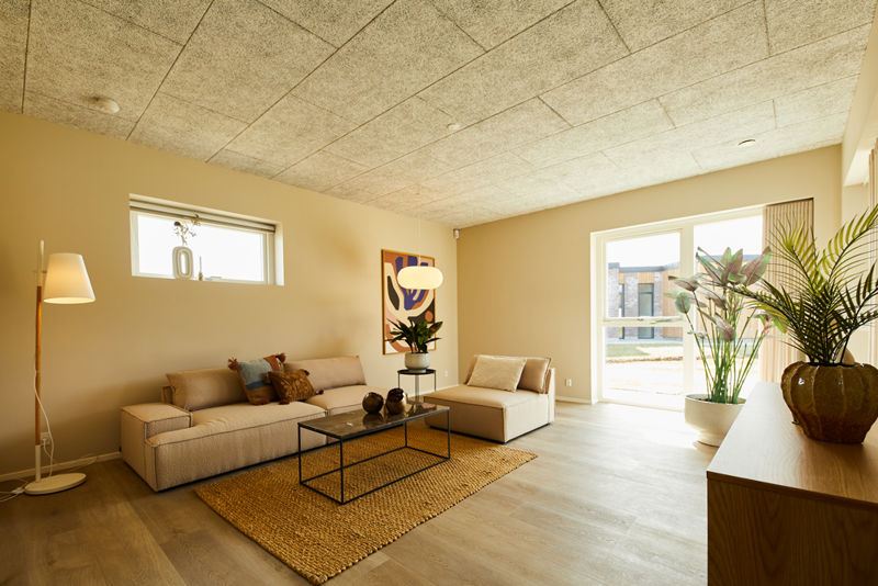 Stue fra eurodan-huse med beige sofa, matchende chaiselong, sort metalsofabord, stort vævet gulvtæppe, høj gulvlampe, grønne planter og stort vinduesparti med dagslys – inspiration til nybyggeri, husbyggeri og indbydende husdrømme.