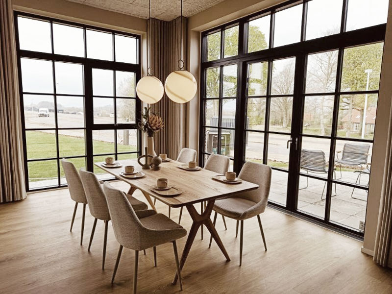 Spiseplads i køkken-alrum fra eurodan-huse med lyst træbord, polstrede spisebordsstole, dækket bord, runde pendellamper og store sorte vinduespartier med udsyn til terrasse – inspiration til nybyggeri, husbyggeri og moderne husdrømme.