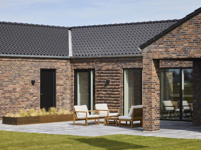 Flisebelagt terrasse ved moderne H-hus med rødbrun murstensfacade, mørkt tegltag, store vinduespartier og loungemøbler – inspiration til nybyg og husdrømme.