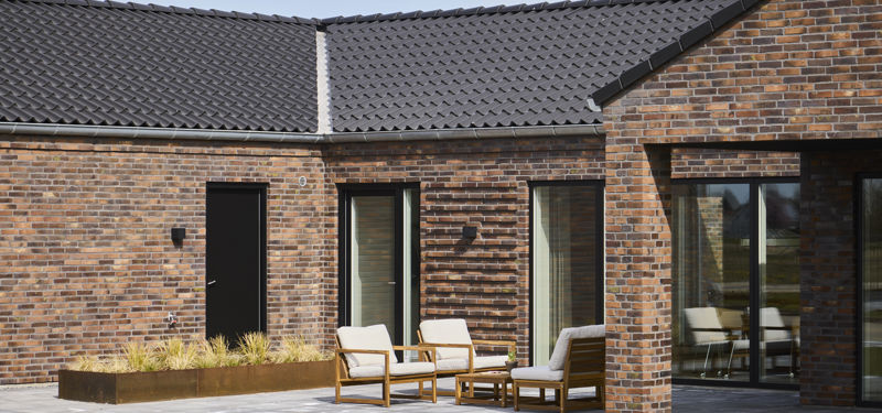Flisebelagt terrasse ved moderne H-hus med rødbrun murstensfacade, mørkt tegltag, store vinduespartier og loungemøbler – inspiration til nybyg og husdrømme.