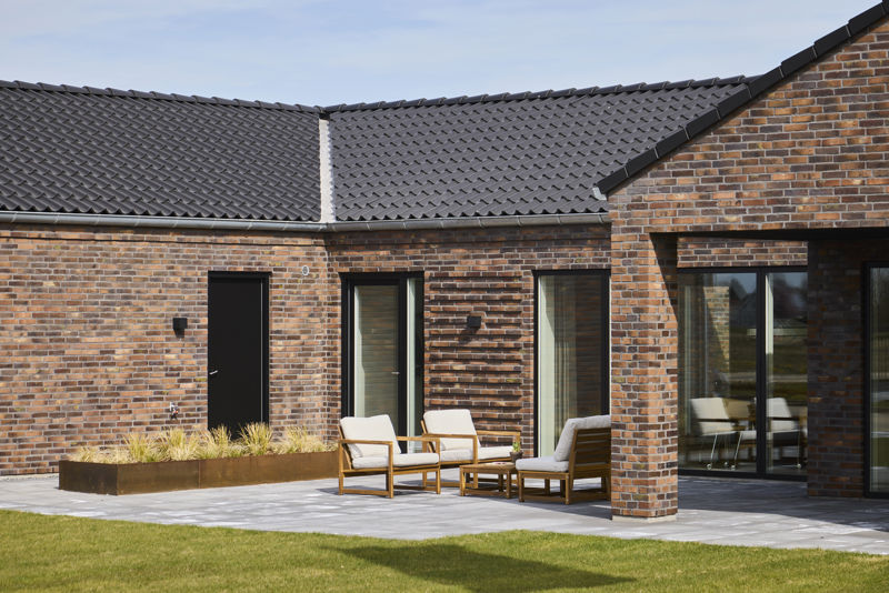 Flisebelagt terrasse ved moderne H-hus med rødbrun murstensfacade, mørkt tegltag, store vinduespartier og loungemøbler – inspiration til nybyg og husdrømme.