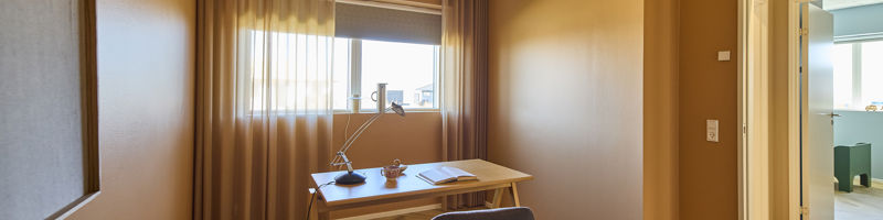 Kontor fra eurodan-huse med træskrivebord, polstret kontorstol, arkitektlampe, tæppe, brune vægge, gardiner og vindue med naturligt lys – inspiration til roligt hjemmekontor i nybyggeri og funktionelle husdrømme.