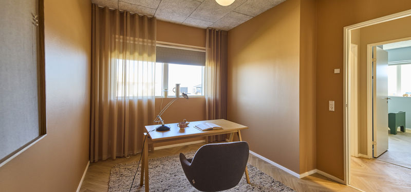 Kontor fra eurodan-huse med træskrivebord, polstret kontorstol, arkitektlampe, tæppe, brune vægge, gardiner og vindue med naturligt lys – inspiration til roligt hjemmekontor i nybyggeri og funktionelle husdrømme.