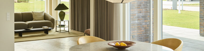 Indbydende spiseplads med rundt spisebord, polstrede stole, stor pendellampe, sildebensgulv og brede vinduespartier med kig til stuen – moderne inspiration til nybyg og husdrømme.