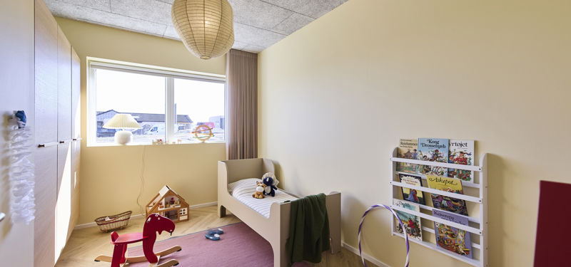 Lyst børneværelse med seng, gyngehest, væghængt bogreol, rosa gulvtæppe, indbyggede skabe og stort vindue – hyggelig inspiration til familiens nybyg og husdrømme.