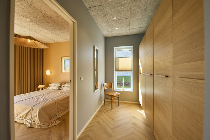 Walk-in-område ved soveværelse fra eurodan-huse med høj skabsvæg i lyst træ, smalle greb, sildebensgulv, lille træstol, vindue med rullegardin og kig ind til seng med gardiner og lampe – inspiration til elegant garderobeplads i nybygget hus og gennemtænkt husbyggeri.