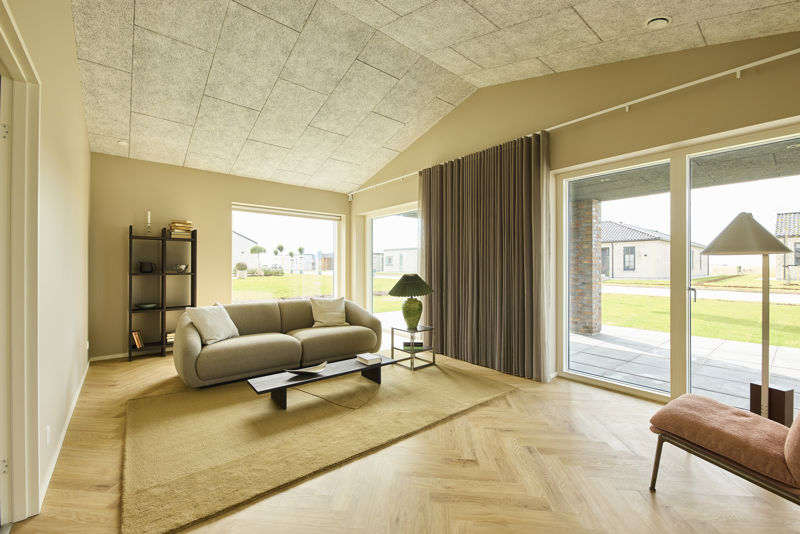 Stue fra eurodan-huse med buet lys sofa, sort lavt sofabord, stor grøn bordlampe, høje gardiner, bred terrassedør, stort vindue og akustikloft med skrå loftflade – inspiration til nybyggeri, husbyggeri og moderne stueindretning.