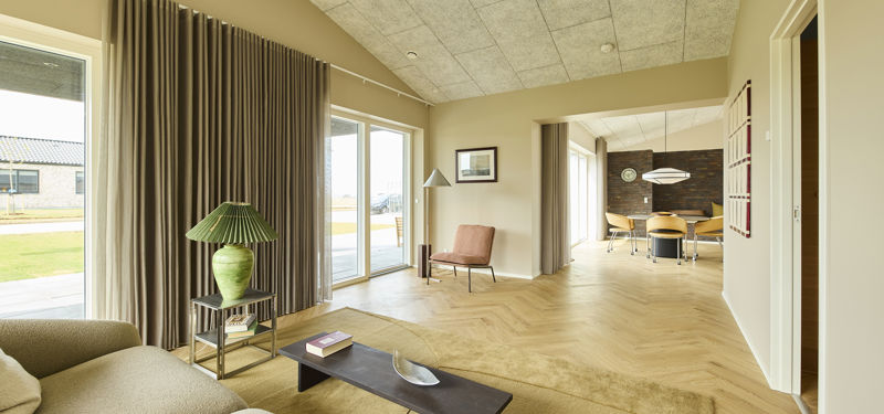 Lys stue med sofa, mørkt sofabord, sildebensgulv, lange gardiner og store vinduespartier samt åben passage til spiseområdet – inspiration til moderne nybyg og husdrømme.