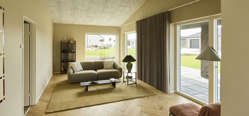 Lys stue med sofa, loungestol, sildebensgulv, lange gardiner og store vinduespartier med udgang til terrassen – indbydende inspiration til moderne nybyg og husdrømme.