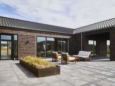 Flisebelagt terrasse fra eurodan-huse med muret facade i varme nuancer, sort tegltag, store vinduespartier, overdækket hjørne, loungemøbler og plantebed med høje græsser – inspiration til terrasse i nybyggeri, husbyggeri og moderne husdrømme.