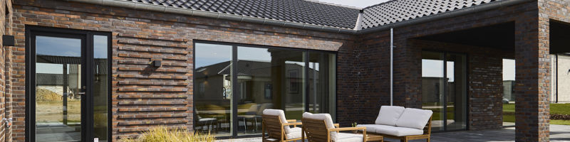 Flisebelagt terrasse fra eurodan-huse med muret facade i varme nuancer, sort tegltag, store vinduespartier, overdækket hjørne, loungemøbler og plantebed med høje græsser – inspiration til terrasse i nybyggeri, husbyggeri og moderne husdrømme.