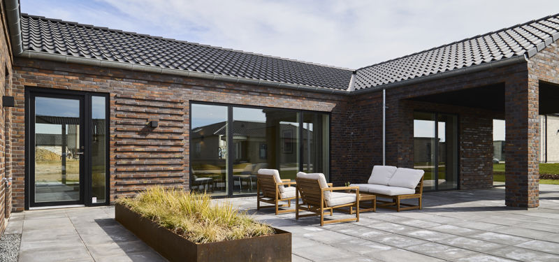 Flisebelagt terrasse fra eurodan-huse med muret facade i varme nuancer, sort tegltag, store vinduespartier, overdækket hjørne, loungemøbler og plantebed med høje græsser – inspiration til terrasse i nybyggeri, husbyggeri og moderne husdrømme.