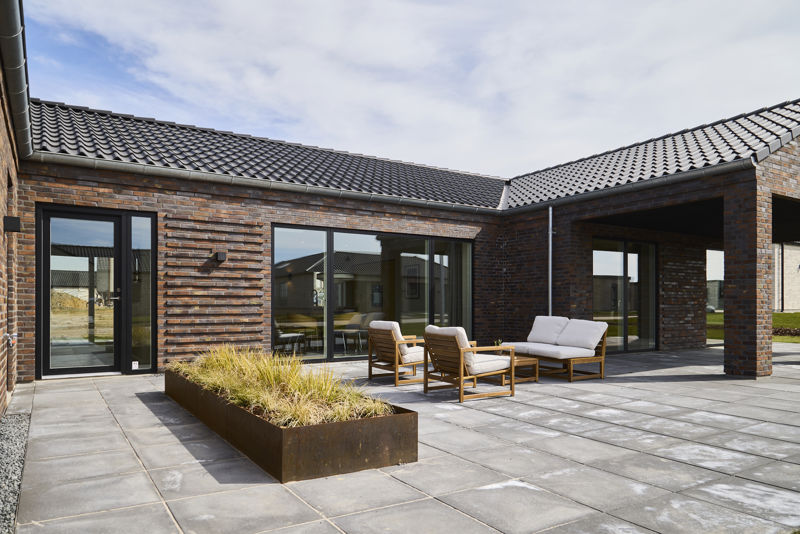 Flisebelagt terrasse fra eurodan-huse med muret facade i varme nuancer, sort tegltag, store vinduespartier, overdækket hjørne, loungemøbler og plantebed med høje græsser – inspiration til terrasse i nybyggeri, husbyggeri og moderne husdrømme.