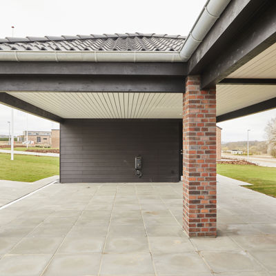 Carport set indefra med røde murstenssøjler, mørke træbjælker, lyst loft, store grå fliser og ladestander på bagvæggen – inspiration til elbilvenlig carport, moderne nybyggeri og smarte husdrømme fra eurodan-huse.