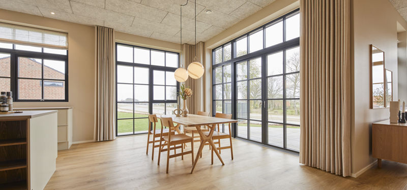 Køkken-alrum fra eurodan-huse med spiseplads ved store sorte vinduespartier, lyse gardiner fra loft til gulv, træbord, træstole, runde pendellamper og udsyn til terrasse – konkret inspiration til nybyggeri og husdrømme med naturligt lys.