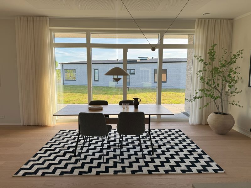 Spiseplads fra eurodan-huse med stort lyst vinduesparti bag spisebordet, hvide gardiner, sort-hvidt zigzag-tæppe, mørke spisebordsstole, pendellampe og stor plante i krukke – inspiration til nybyggeri, husbyggeri og lyse opholdsrum.