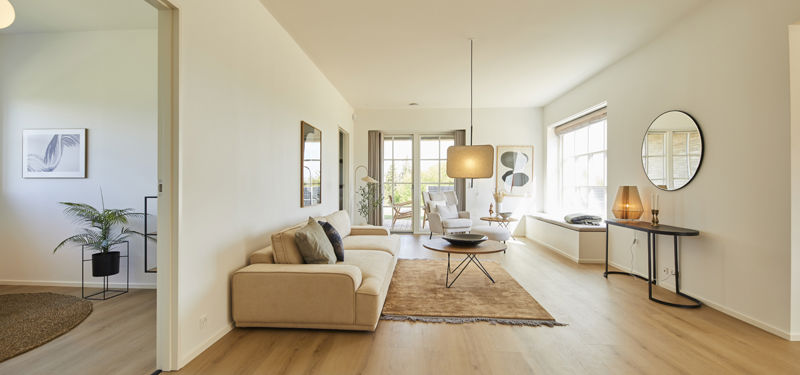Lang stue fra eurodan-huse med beige sofa, rundt sofabord, stort tæppe, væghængt spejl, konsolbord med lampe, indbygget vinduesniche og åbent kig til ekstra opholdsrum – inspiration til palæstil, nybyggeri og lyse husdrømme.