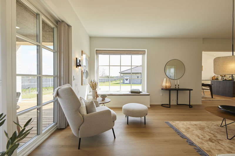 Lyst stuehjørne fra eurodan-huse med beige lænestol, rund puf, stort vindue, bredt sansevindue med sort hynde, rundt spejl på væggen og kig mod køkken – inspiration til nybyggeri, husbyggeri og smarte opholdsrum i nybygget hus.