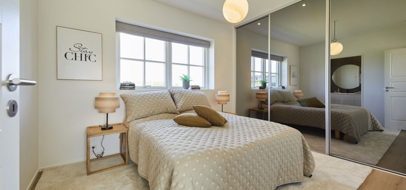 Soveværelse fra eurodan-huse med beige seng og prikket sengetæppe, små natborde med lamper, bredt vindue med rullegardin, stor garderobe med spejlskydelåger og blødt gulvtæppe – inspiration til nybyggeri, husbyggeri og soveværelse med ekstra opbevaring.