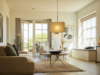 Lys stue fra eurodan-huse med beige sofa, loungestol, rundt sofabord, stor firkantet pendellampe, sansevindue med hynde, store vinduer og bløde gardiner mod terrassen – inspiration til nybyggeri, husbyggeri og hyggelige opholdsrum.