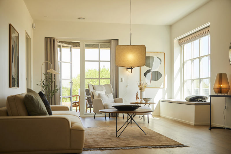 Lys stue fra eurodan-huse med beige sofa, loungestol, rundt sofabord, stor firkantet pendellampe, sansevindue med hynde, store vinduer og bløde gardiner mod terrassen – inspiration til nybyggeri, husbyggeri og hyggelige opholdsrum.