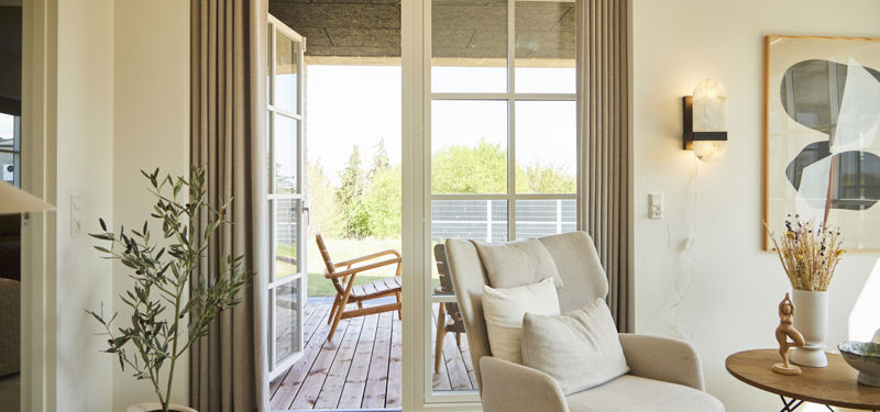 Udgang til terrasse fra eurodan-huse med hvid sprosset terrassedør, højt vinduesparti, lange beige gardiner, lys lænestol og kig tdækket udil overerum – inspiration til nybyggeri med godt lysindfald og sammenhæng mellem bolig og terrasse.