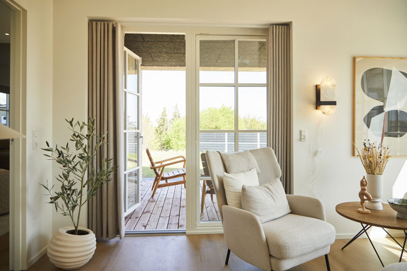 Udgang til terrasse fra eurodan-huse med hvid sprosset terrassedør, højt vinduesparti, lange beige gardiner, lys lænestol og kig tdækket udil overerum – inspiration til nybyggeri med godt lysindfald og sammenhæng mellem bolig og terrasse.
