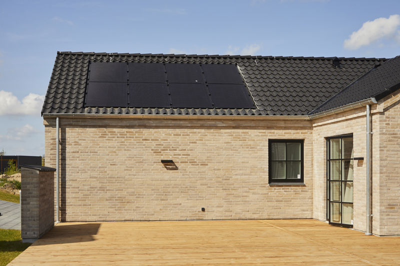 Træterrasse fra eurodan-huse langs lys murstensfacade med sorte vinduesrammer, mørkt tegltag, synlige solceller og bredt terrasseareal foran huset – inspiration til nybygget hus med terrasse, solceller og praktisk udeliv.